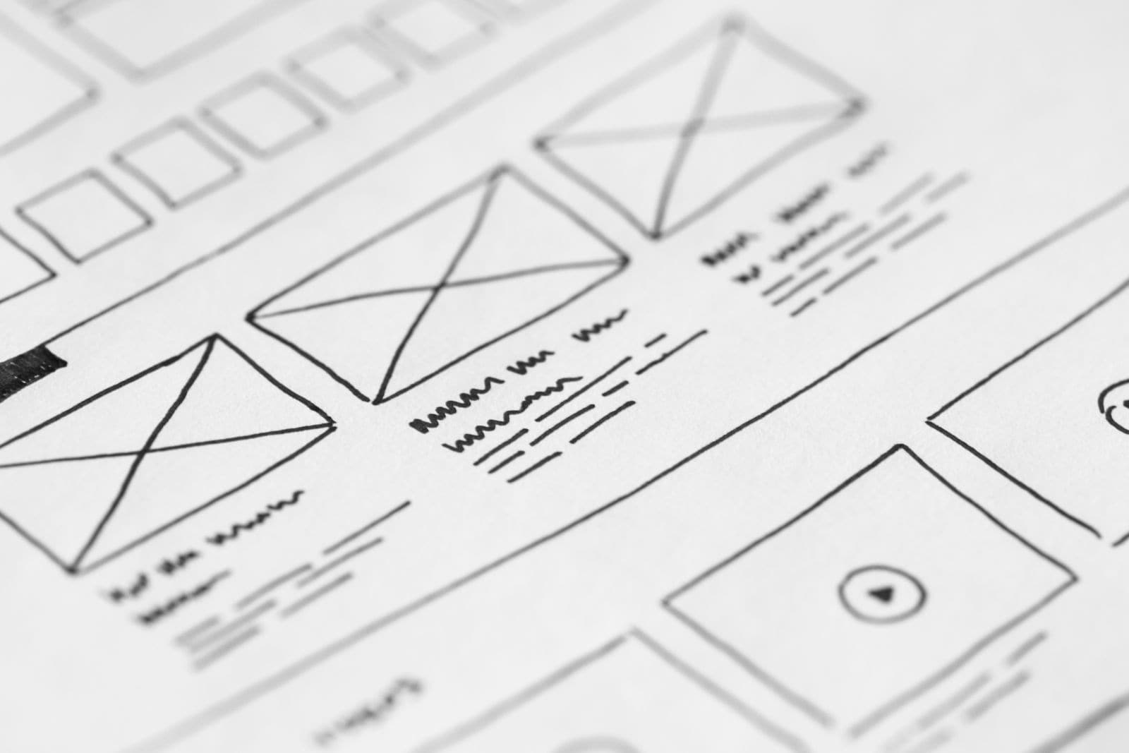 A hand-drawn website wireframe sketch on paper showing header, image blocks and body-text placeholders